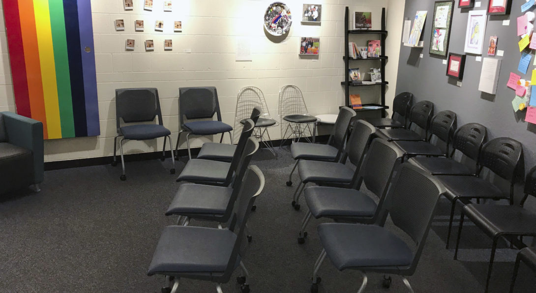 Color image of an empty space. There is multiple art pieces on the wall from the Queer State of Mind Exhibition. Many chairs are against the wall and set up in rows facing the same direction.
