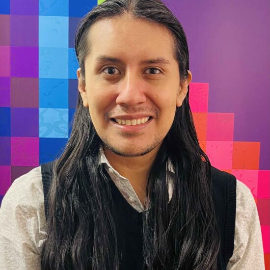 Moisés is wearing a black vest, and a white shirt. Moisés has long straight black hair, and is standing in front of a purple background with multicolored rectangles.