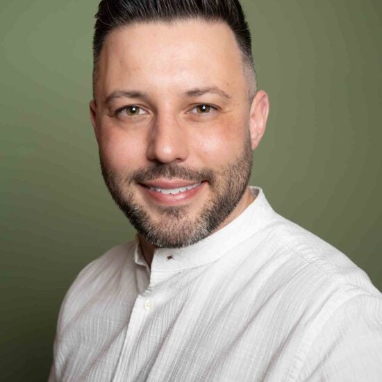 Nic behind a green olive color backgroudn wearing a white shirt, smiling and black stubbly facial hair.