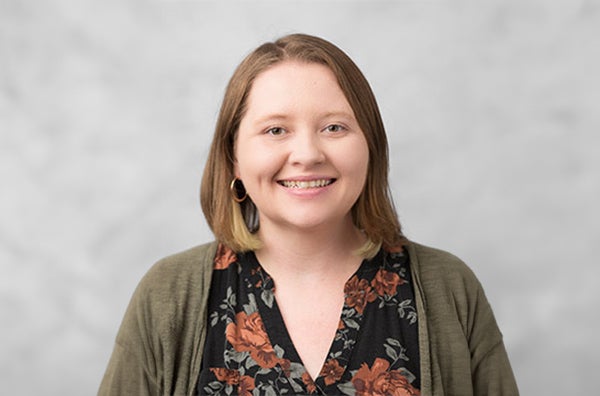 Amanda wearing a floral blouse with dark green sweater, smiling, in front of grey/white backdrop.