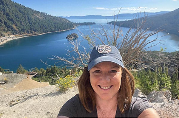 Debra smiling with a cap on in front of a lake.