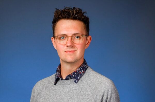 James is wearing glasses and a patterned shirt underneath a grey sweater in front of a blue background.