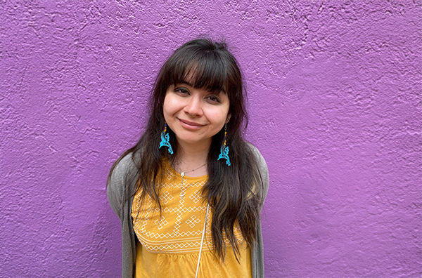 Mariela smiling wearing blue earrings, yellow top, and grey sweater in front of a purple wall.