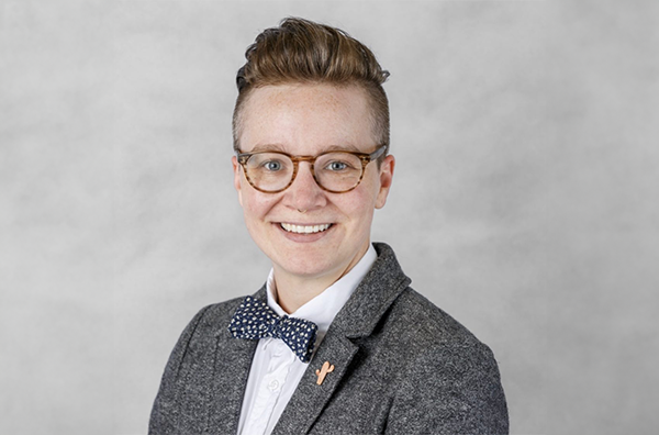 Mimi is wearing glasses, a blue spotted bowtie, a white shirt, and a grey blazer while smiling in front of a light grey background.