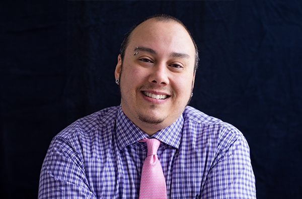 Nat smiling wearing a checkered lavender shirt and pink tie in front of a black background.