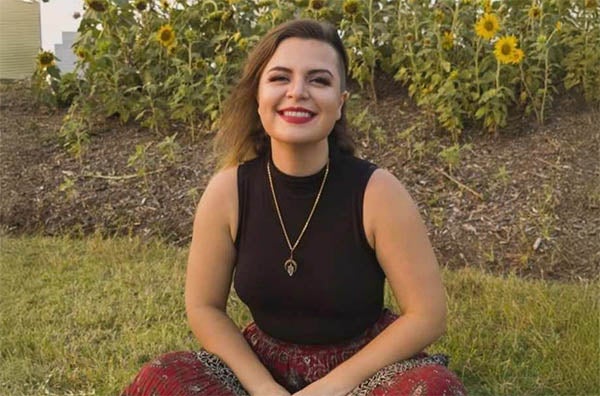 Samantha smiling, wearing a black top, chain around her neck, red pants, sitting on grass with sunflowers behind her.