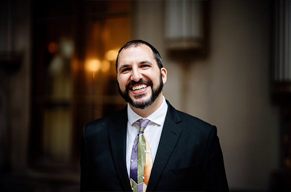 Samus smiling wearing a colorful tie, white shirt, and black blazer in front of a building's facade.