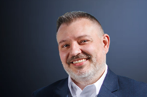 Shawn smiling wearing a white shirt and navy blazer in front of a dark blue backdrop.