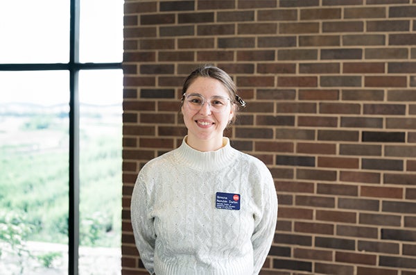 Simone smiling, wearing glasses, white turtleneck, in front of a brick wall.